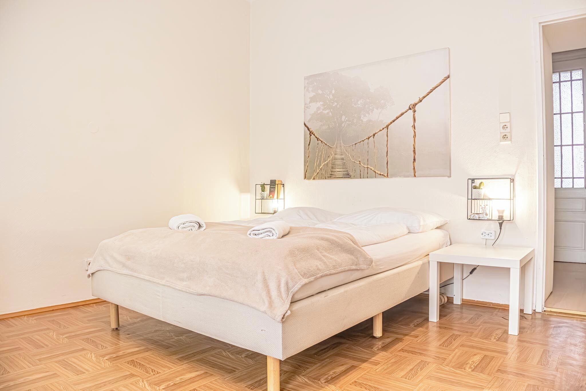 Un lit bien fait, recouvert d’une couverture beige, une table de chevet avec une lampe et, au mur, une image représentant un pont de corde.
