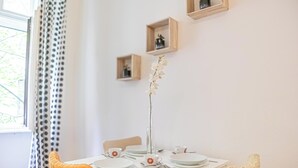 Une table à manger est dressée avec des assiettes, des tasses et un vase rempli de fleurs. Il y a deux chaises, dont une est dotée d’un coussin texturé.