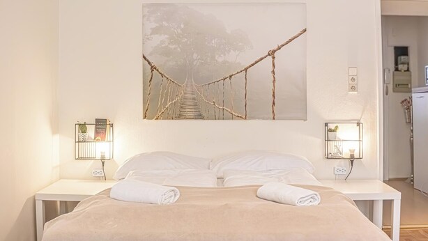 Une chambre à coucher comprenant un lit, deux tables de chevet, un tableau représentant un pont accroché au mur, et deux lampes fixées au mur.