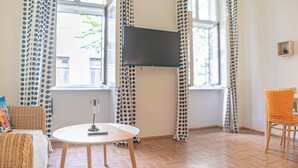 Une pièce avec une table ronde, un canapé en osier, un téléviseur fixé au mur et une fenêtre avec des rideaux.