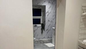 A narrow hallway with a wooden floor leading to a bathroom with a marble wall and a window.
