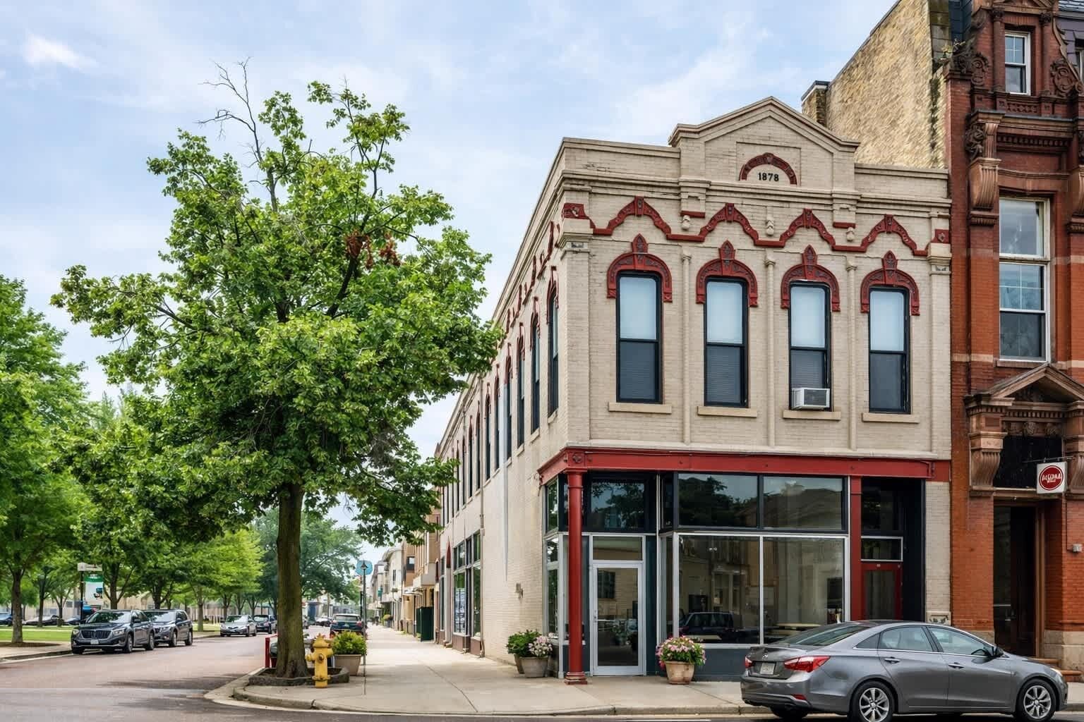 A two-story building with a large tree in front, a fire hydrant, and parked cars on the street.