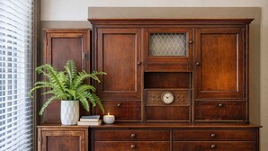 A wooden cabinet with drawers and a clock, a potted fern, and a candle on a windowsill. - Stylish Downtown Loft – Free Parking & City Views! (Oshkosh)