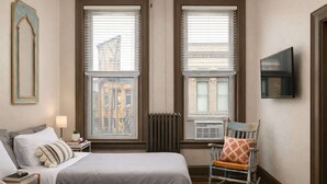 A bedroom with two beds, a rocking chair, a TV, and a view of buildings through the windows. - Stylish Downtown Loft – Free Parking & City Views! (Oshkosh)