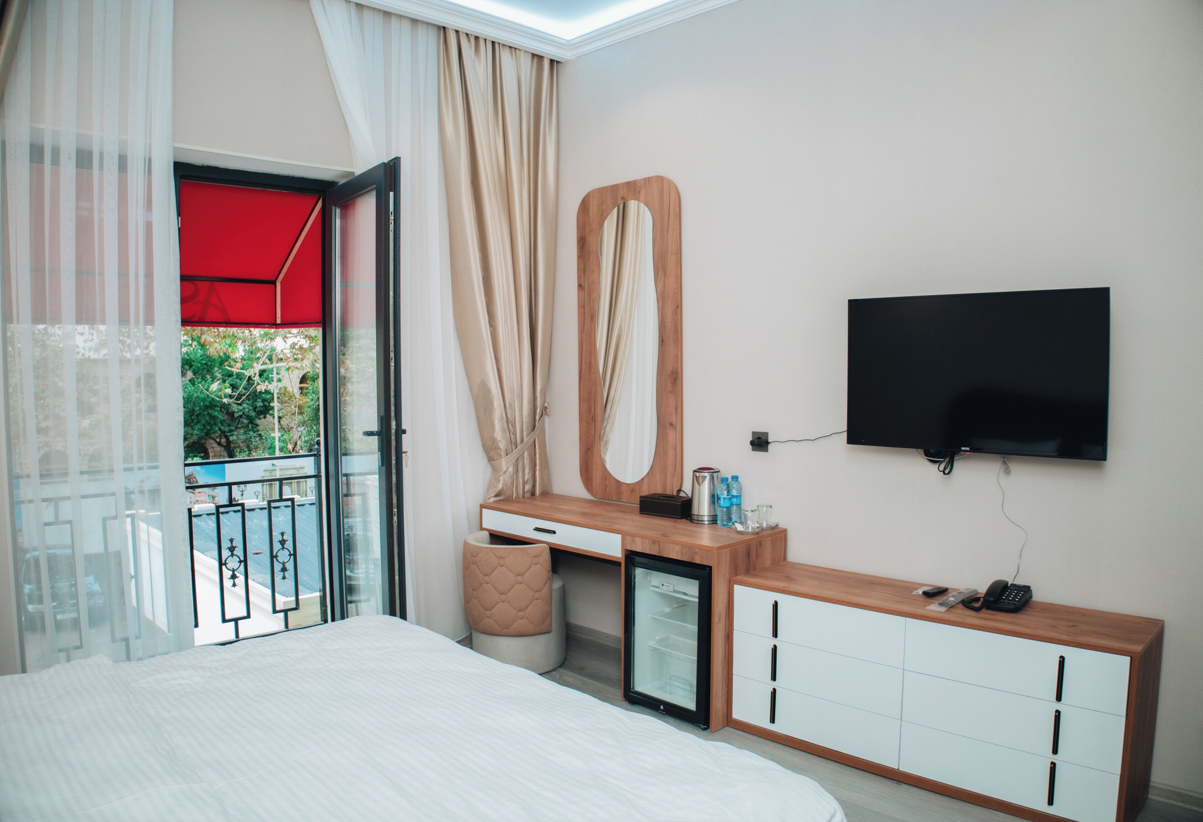 A hotel room with a bed, a desk, a small refrigerator, a TV, and a balcony with a red awning.