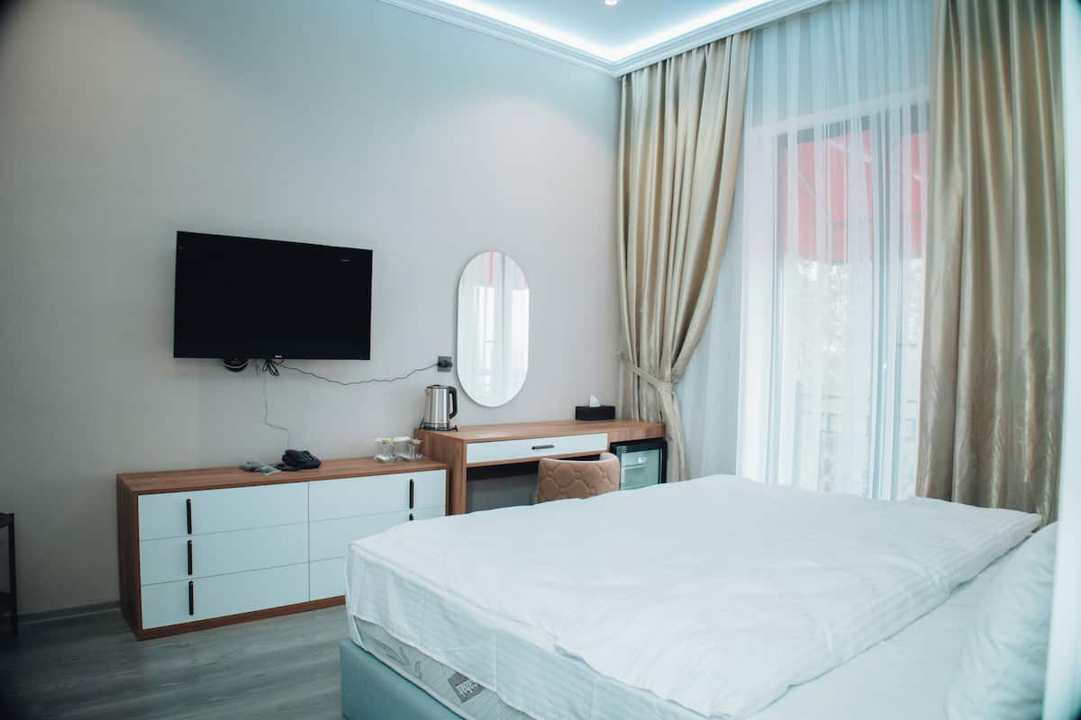 A hotel room with a bed, a desk, a television mounted on the wall, a mirror, and a window with curtains.