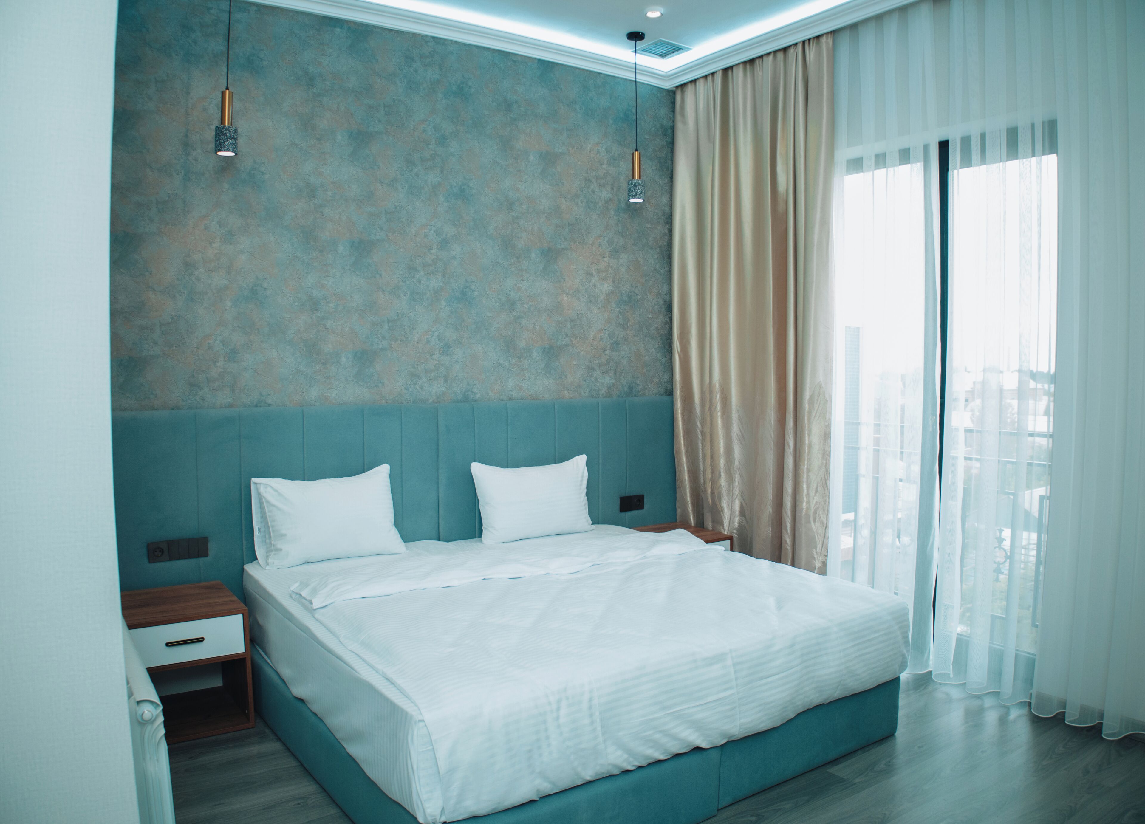 A modern hotel room with a large bed, a nightstand, and a view of the city through sheer curtains.
