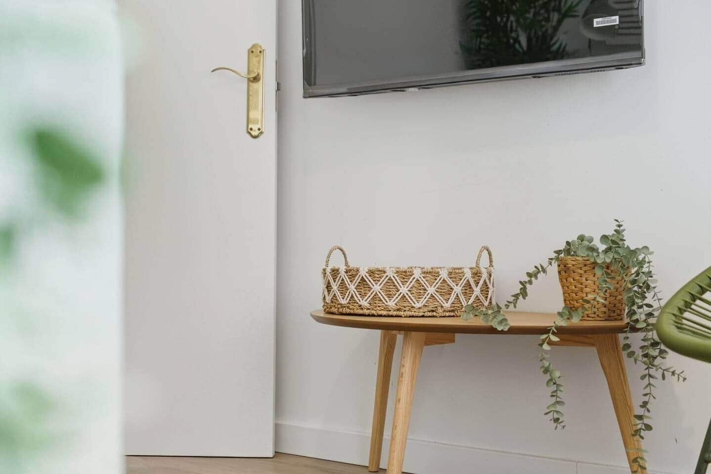 Quarto com porta branca, mesa de madeira com cesta trançada e planta em vaso, além de uma cadeira verde.