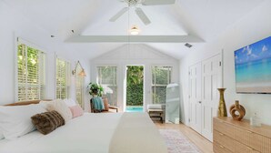 A bedroom with a bed, a ceiling fan, a large window with blinds, a bedside table, a dresser, and a painting on the wall.