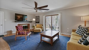 A living room with a ceiling fan, a flat-screen TV, a fireplace, a sofa, a coffee table, and a large window with curtains. - *Pet Friendly* Charming-Single story | Fenced Yard, Outdoor Dining, Near Duke (Durham)