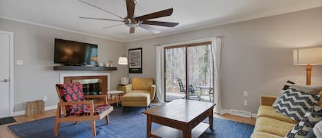A living room with a ceiling fan, a flat-screen TV, a fireplace, a sofa, a coffee table, and a large window with curtains.