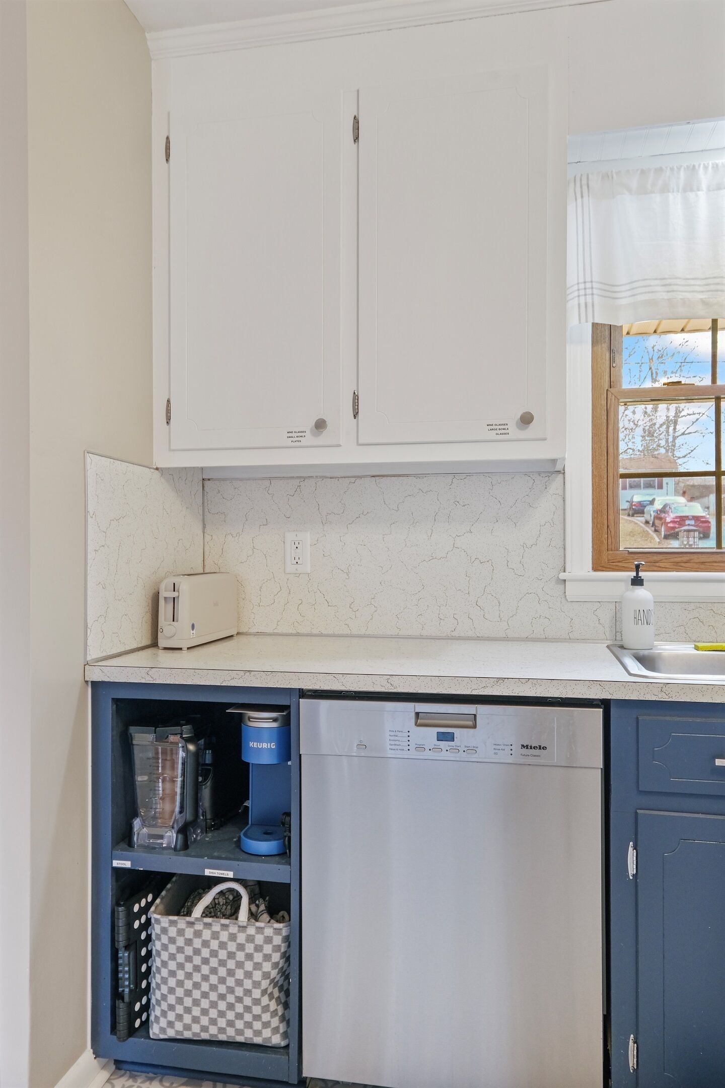 A kitchen with white cabinets, a built-in dishwasher, a coffee maker, and a window with a view of the outside.