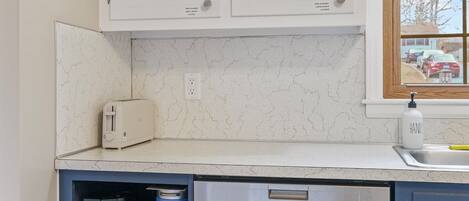 A kitchen with white cabinets, a built-in dishwasher, a coffee maker, and a window with a view of the outside.