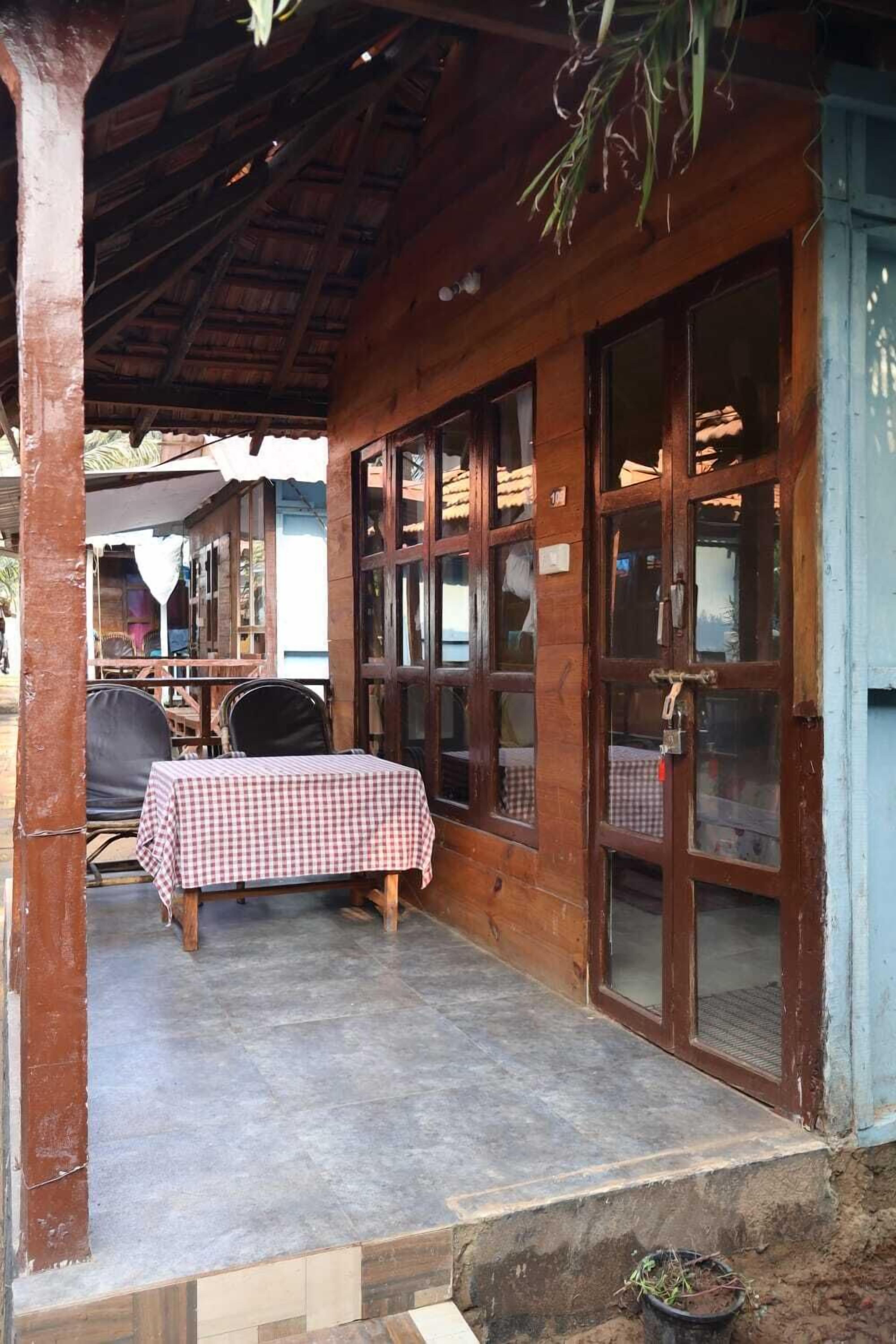 A wooden building with a covered patio area, featuring a table with a checkered tablecloth, chairs, and a potted plant.