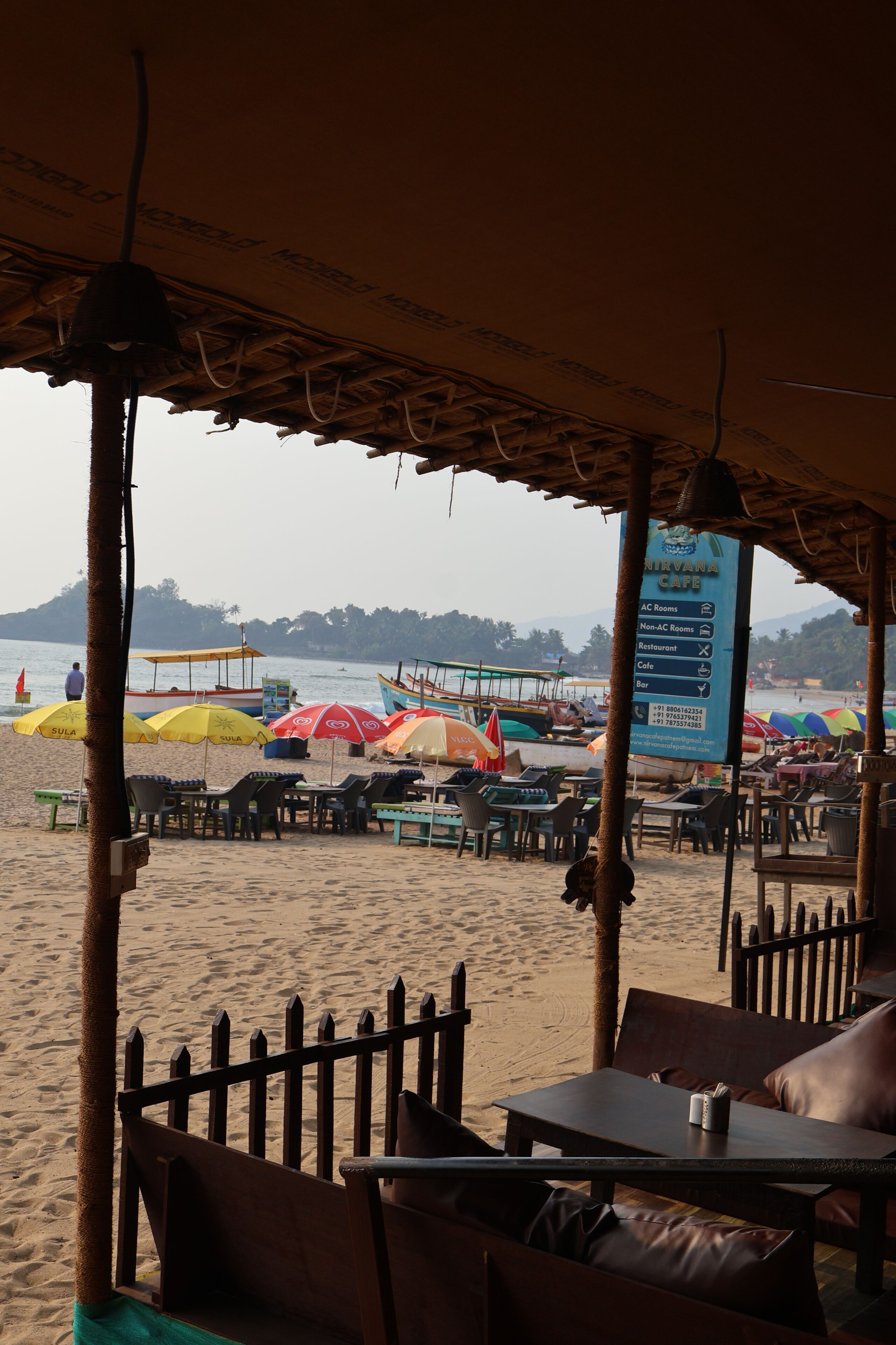A beachside cafe with outdoor seating and umbrellas, overlooking the beach and boats.