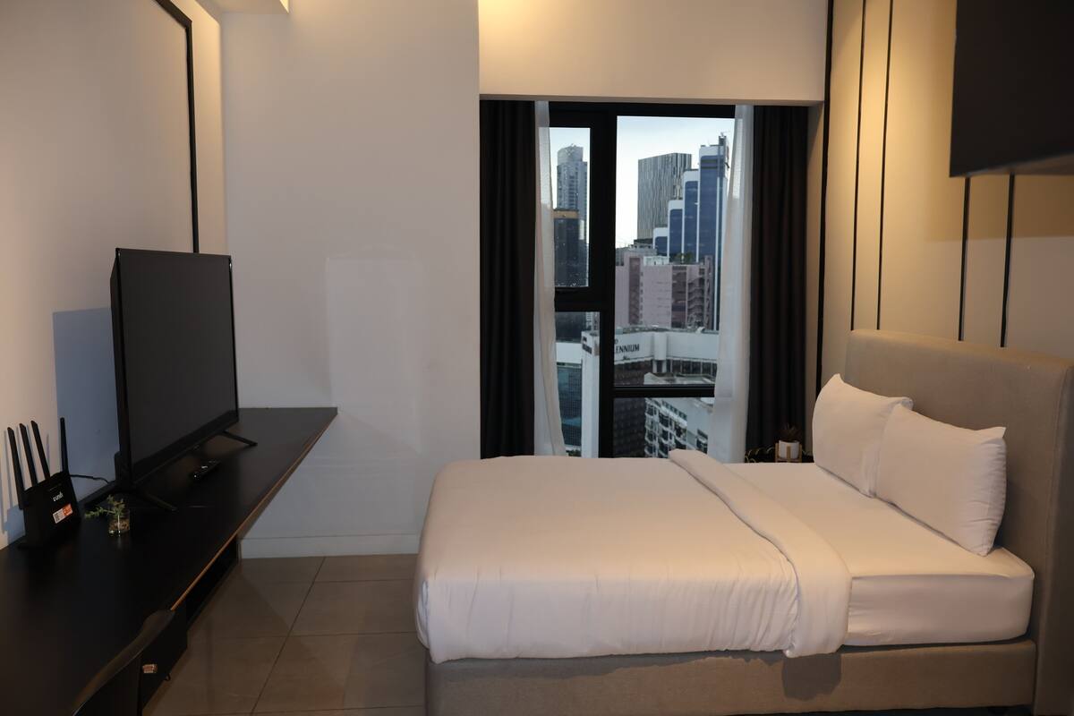 A modern hotel room with a large bed, a flat-screen TV, a desk with a computer, and a view of the cityscape through the window.