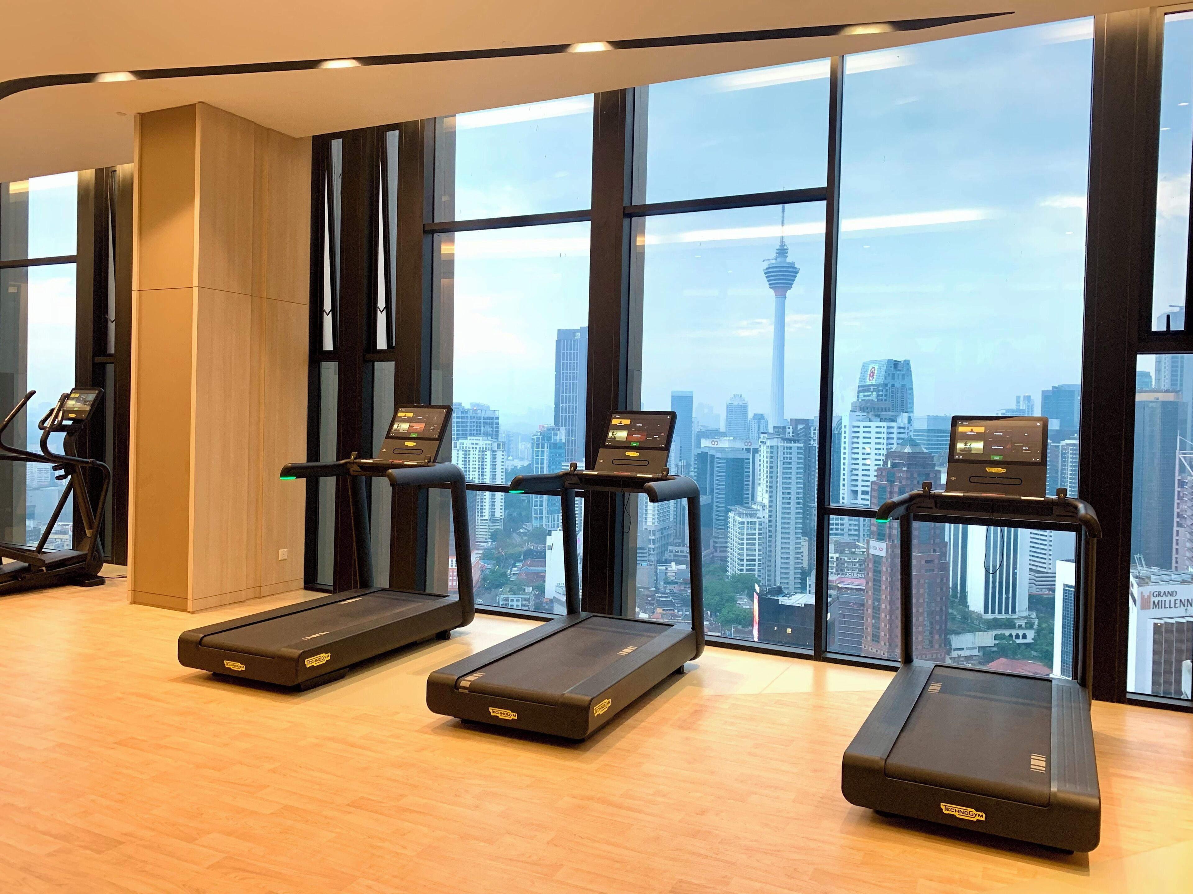 Een fitnessruimte met drie loopbanden, elk met een digitaal scherm, en grote ramen met uitzicht op de stad.