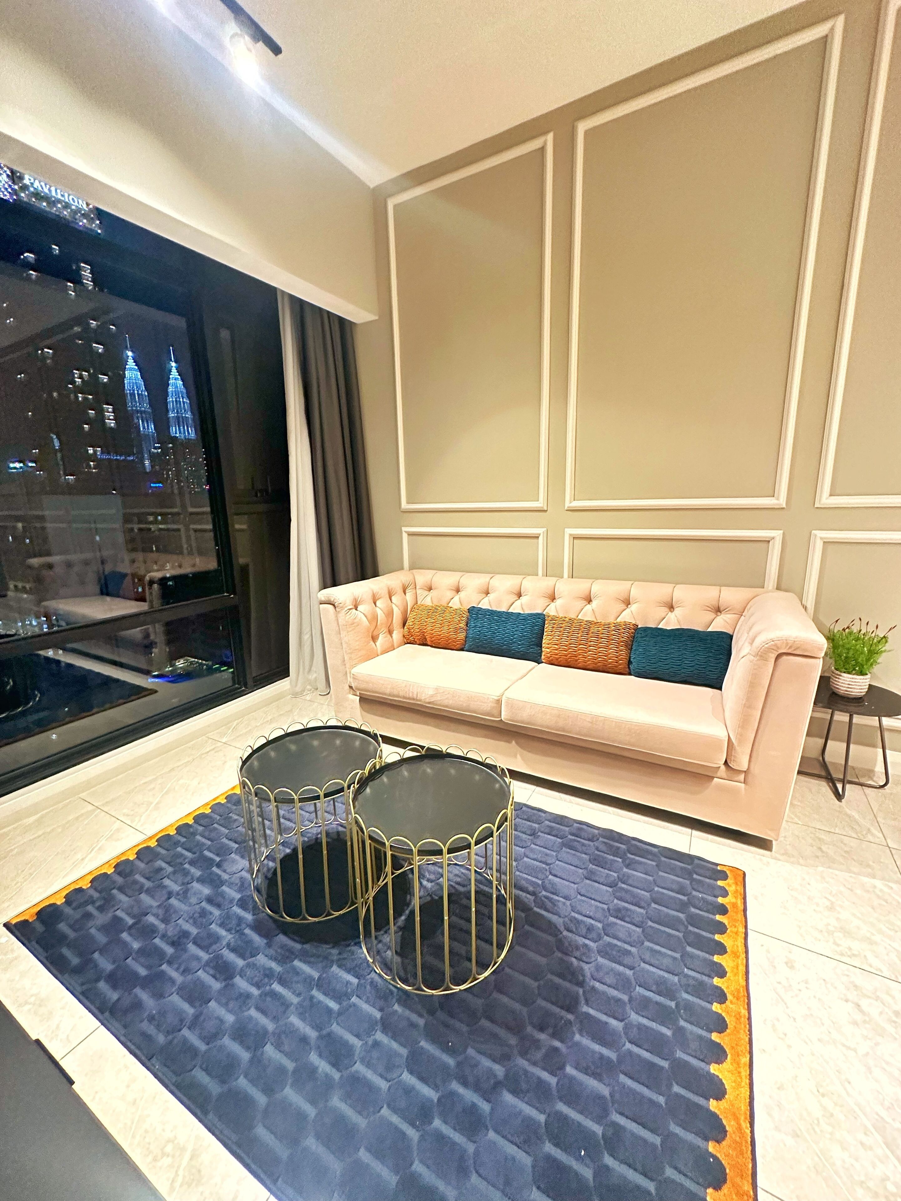 A living room with a beige sofa, two black metal chairs, a blue patterned rug, and a view of a cityscape through a window.