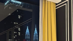 A hotel room with a bed, a window with curtains, and a view of the cityscape including the Petronas Towers.