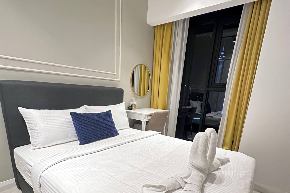 A neatly made bed with a towel, a bedside table, a mirror, and a window with curtains.