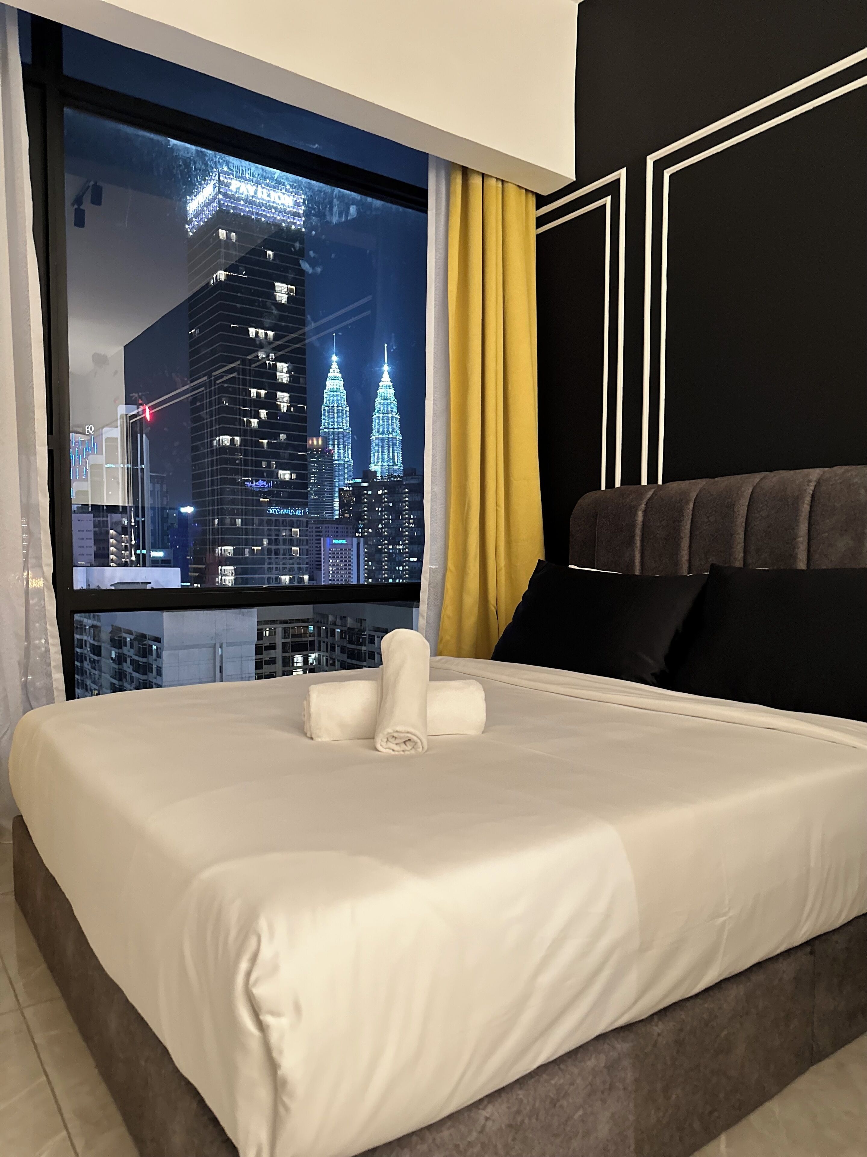 A hotel room with a large window offering a nighttime view of a city skyline, including a prominent building with twin towers. The room features a neatly made bed with white linens and a pair of rolled towels.
