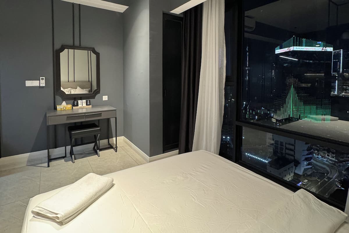 A modern hotel room with a large bed, a desk, a mirror, and a city view through large windows.