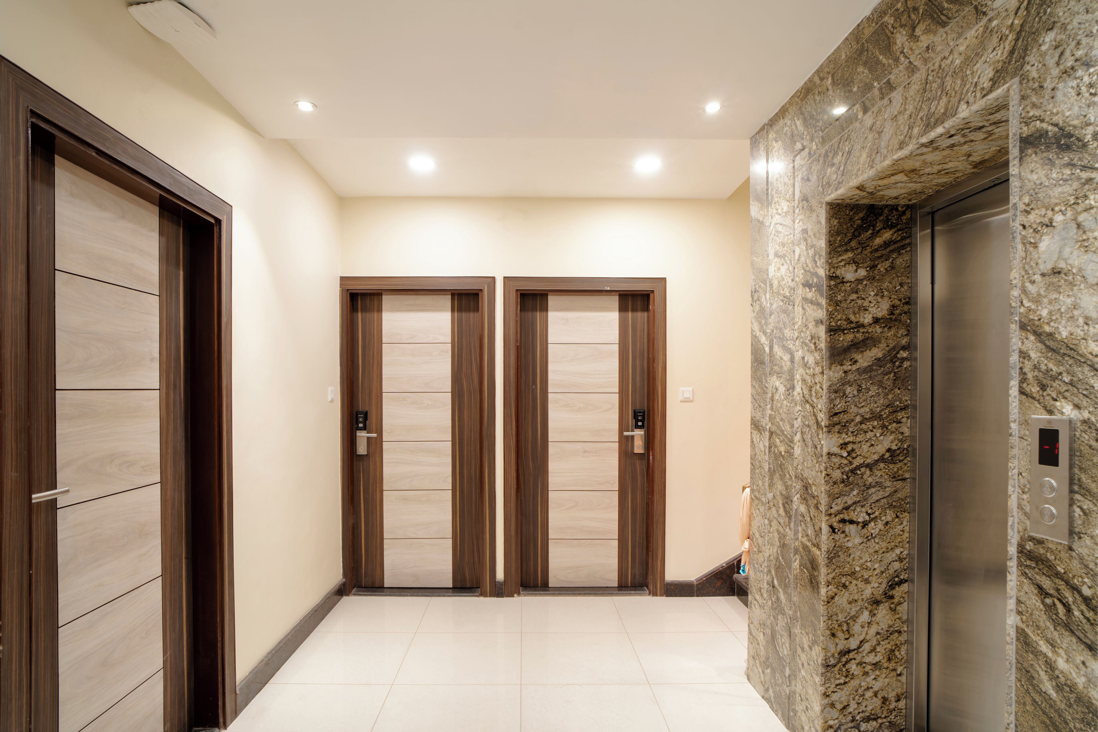 A hallway with two wooden doors and a marble wall with an elevator.