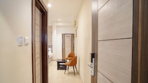 A hotel corridor with a wooden door, a chair, a small table, and a bed in the background. - Omkar International Hotel (VARANASI)
