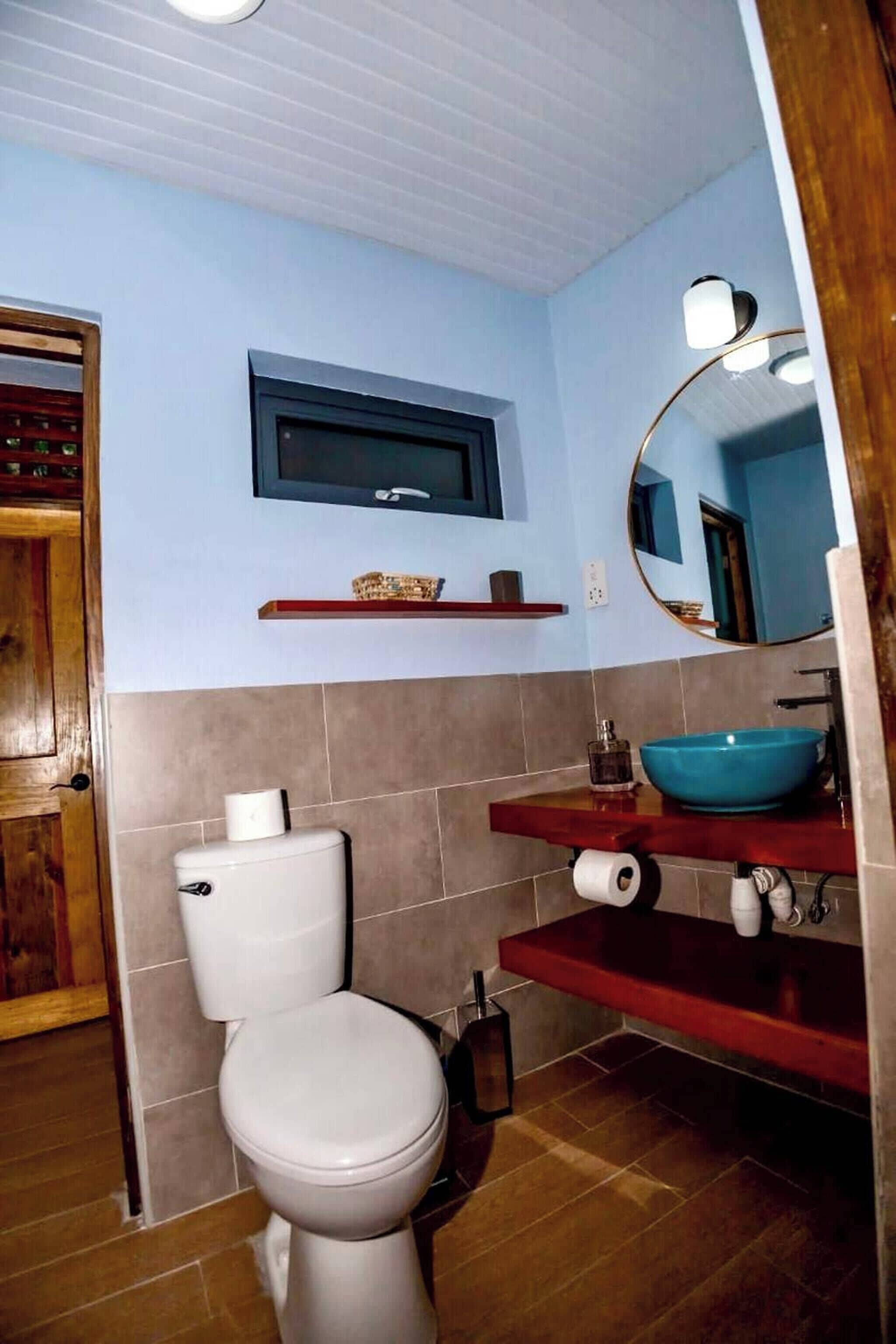 A bathroom with a toilet, a sink with a blue basin, a mirror, and a shelf with toiletries.