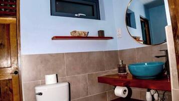 A bathroom with a toilet, a sink with a blue basin, a mirror, and a shelf with toiletries.