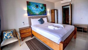 A modern hotel room with a large bed, a painting of a tropical scene, a wooden nightstand, a bench, and a wardrobe. - Hammock CoCo-Nut - by ZenBreak (Morne Fortune)