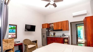 A compact hotel room with a bed, a desk with a chair, a television, a kitchenette, and a ceiling fan.