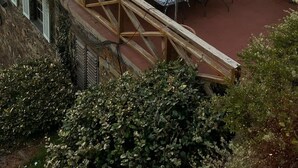 Un edificio rústico de piedra con techo inclinado, zona de estar al aire libre con mesas y sillas, y vista al atardecer de fondo.