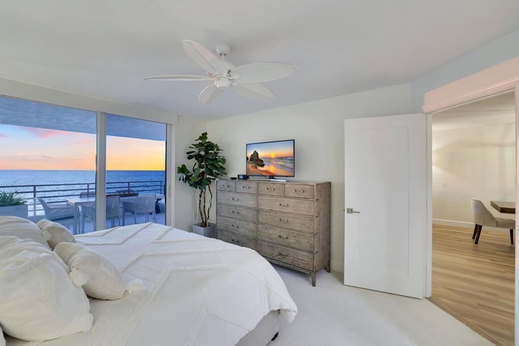Quarto com uma cama grande, um ventilador de teto, cômoda, televisão e vista para o mar através de grandes janelas.