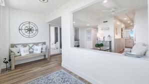 A modern interior with a wooden bench, a compass wall decoration, and a view into a kitchen area. - Beachfront 2BR/2BA Somerset condo with pool, WiFi, and heated pool. (Marco Island)