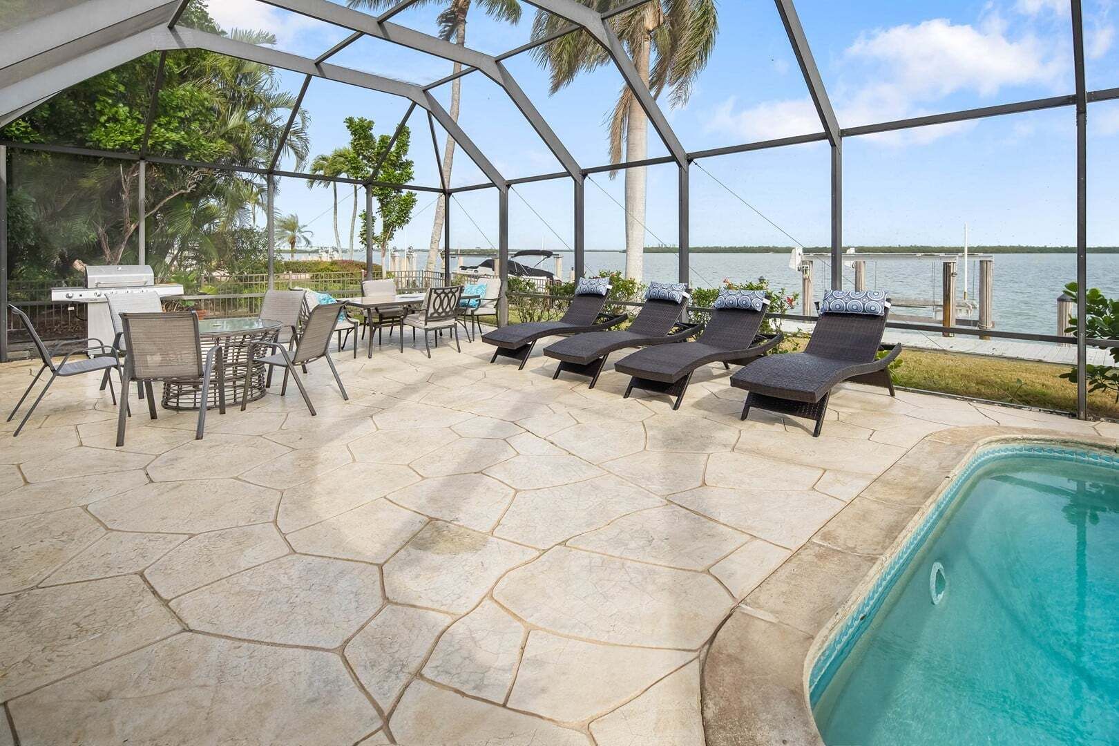 A covered outdoor area with a pool, lounge chairs, a grill, and a table with chairs.