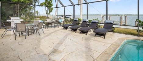A covered outdoor area with a pool, lounge chairs, a grill, and a table with chairs.