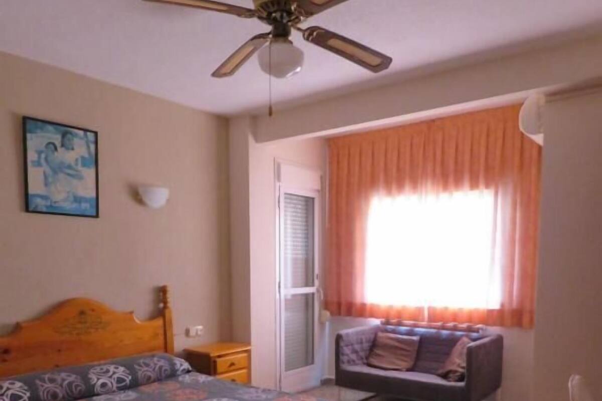 A bedroom with a bed, a desk, a chair, a ceiling fan, a window with curtains, and a door.