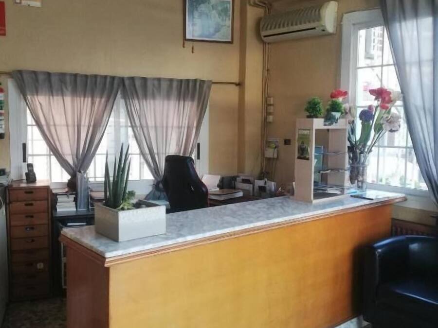 Una zona de recepción con mostrador de madera, ventilador de techo, ventana con cortinas y suelo de baldosas.