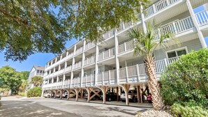 Un hotel de varios pisos con balcones, estacionamiento techado y abundante vegetación.