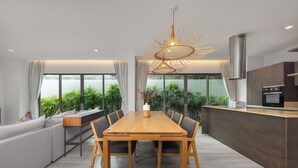 A modern kitchen with a dining area, featuring a wooden table and chairs, a unique hanging light fixture, and a large window with a view of greenery. - Villa Mauri Modern 3BR Private Pool (Rawai)