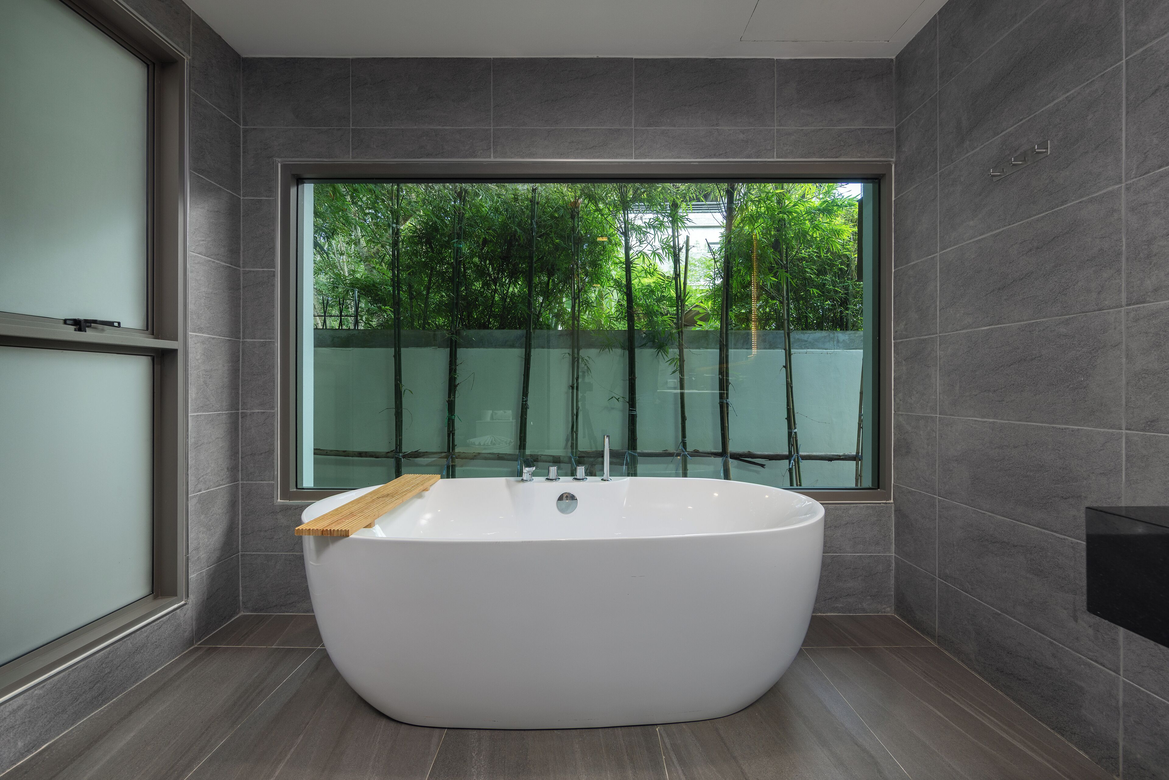 Een moderne badkamer met een vrijstaand bad, een groot raam met uitzicht op groen, en een betegelde vloer.