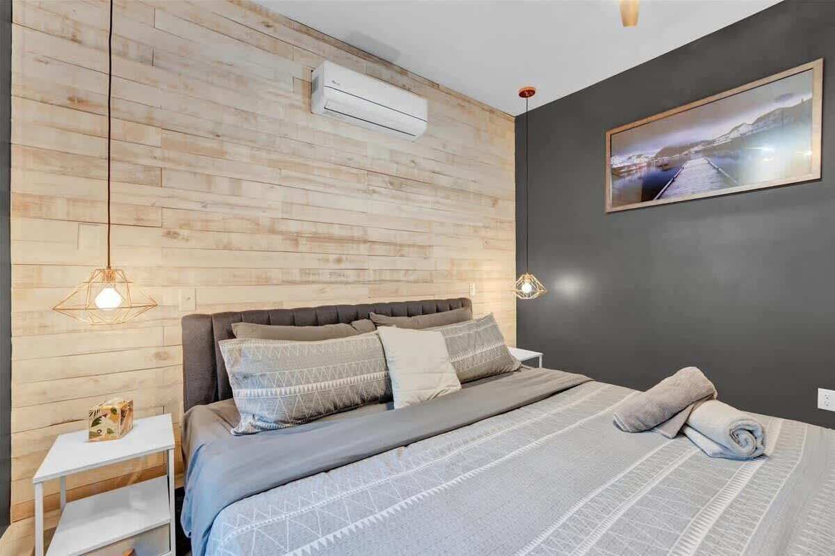Ein modernes Schlafzimmer mit einer Holzvertäfelung, einem Bett mit grauer Bettwäsche, einem weißen Nachttisch und einer Wand-Klimaanlage.