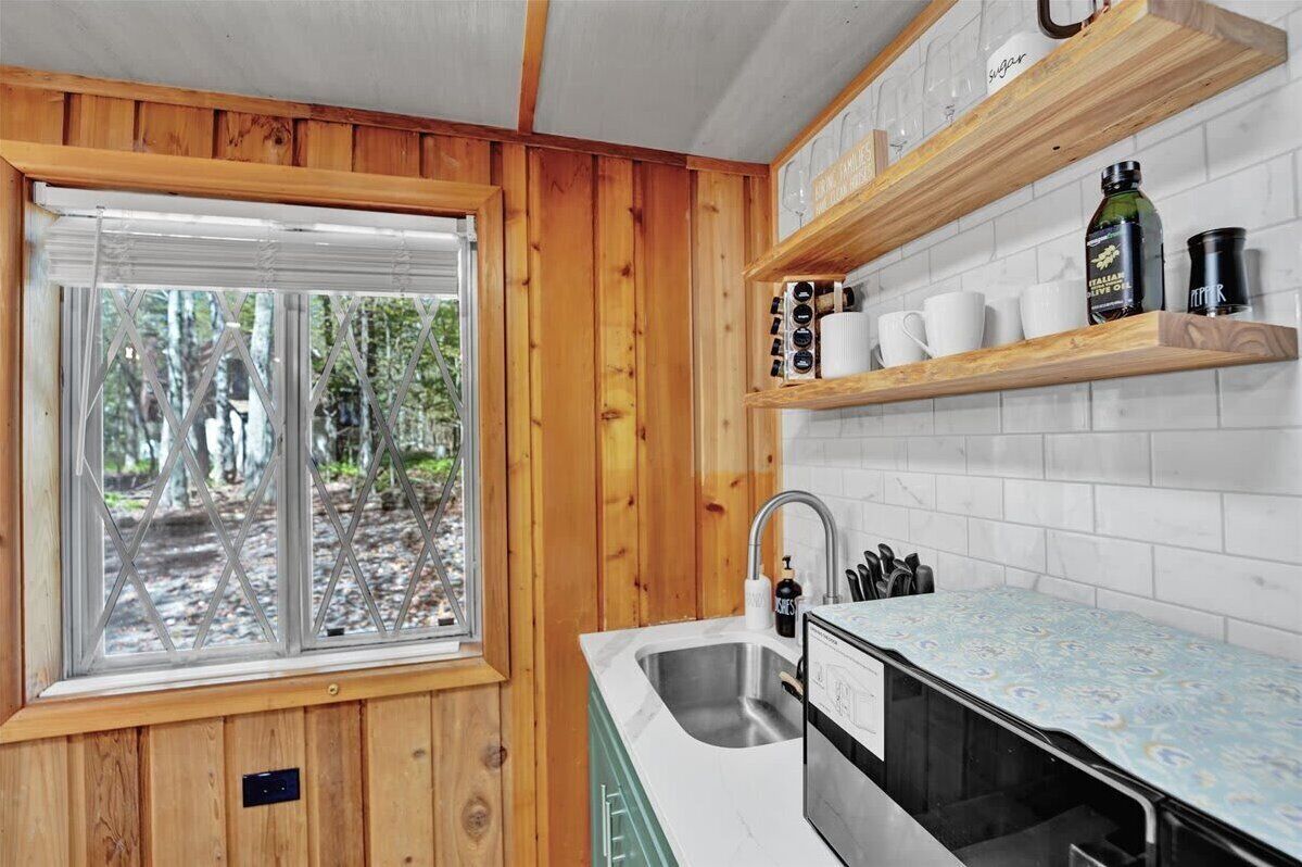 A kitchen with wooden walls, a window with a view of trees, a sink, and shelves with kitchen items.