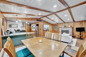 A rustic cabin interior with a wooden dining table, chairs, a kitchen area, a fireplace, and a television.