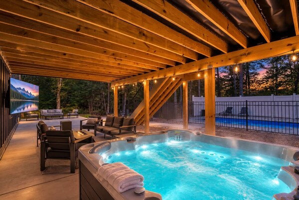 A hot tub under a wooden pergola with outdoor seating and a pool in the background.