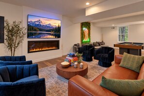 Een moderne woonkamer met een open haard, comfortabele zithoeken, een pooltafel en een grote televisie waarop een berglandschap wordt getoond.