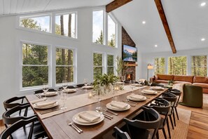 A dining area with a long wooden table set for a meal, surrounded by chairs, with a fireplace and a sofa in the background.