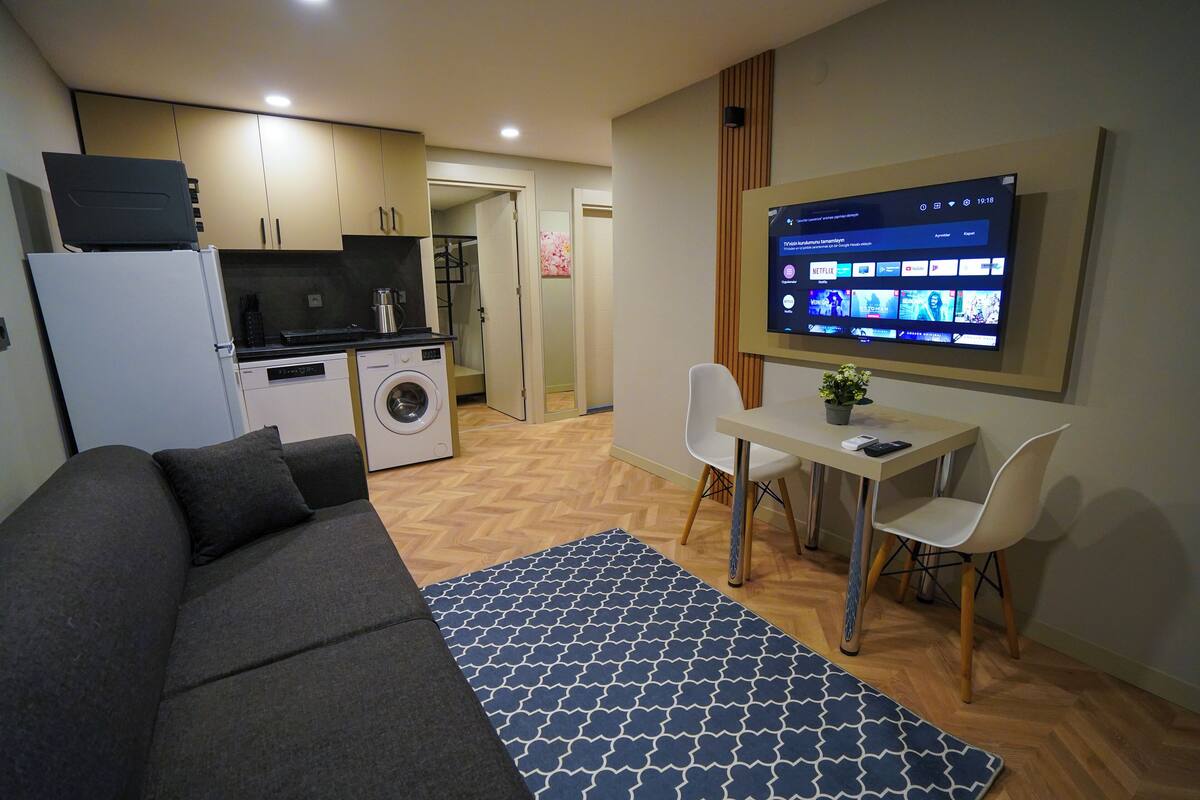 A compact living space with a sofa, a dining table with chairs, a washing machine, and a television mounted on the wall displaying a streaming service interface.