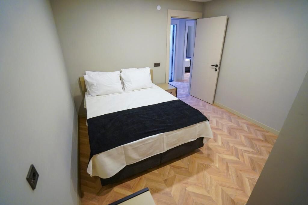 A bedroom with a bed, a wooden herringbone floor, a door, and a light switch on the wall.